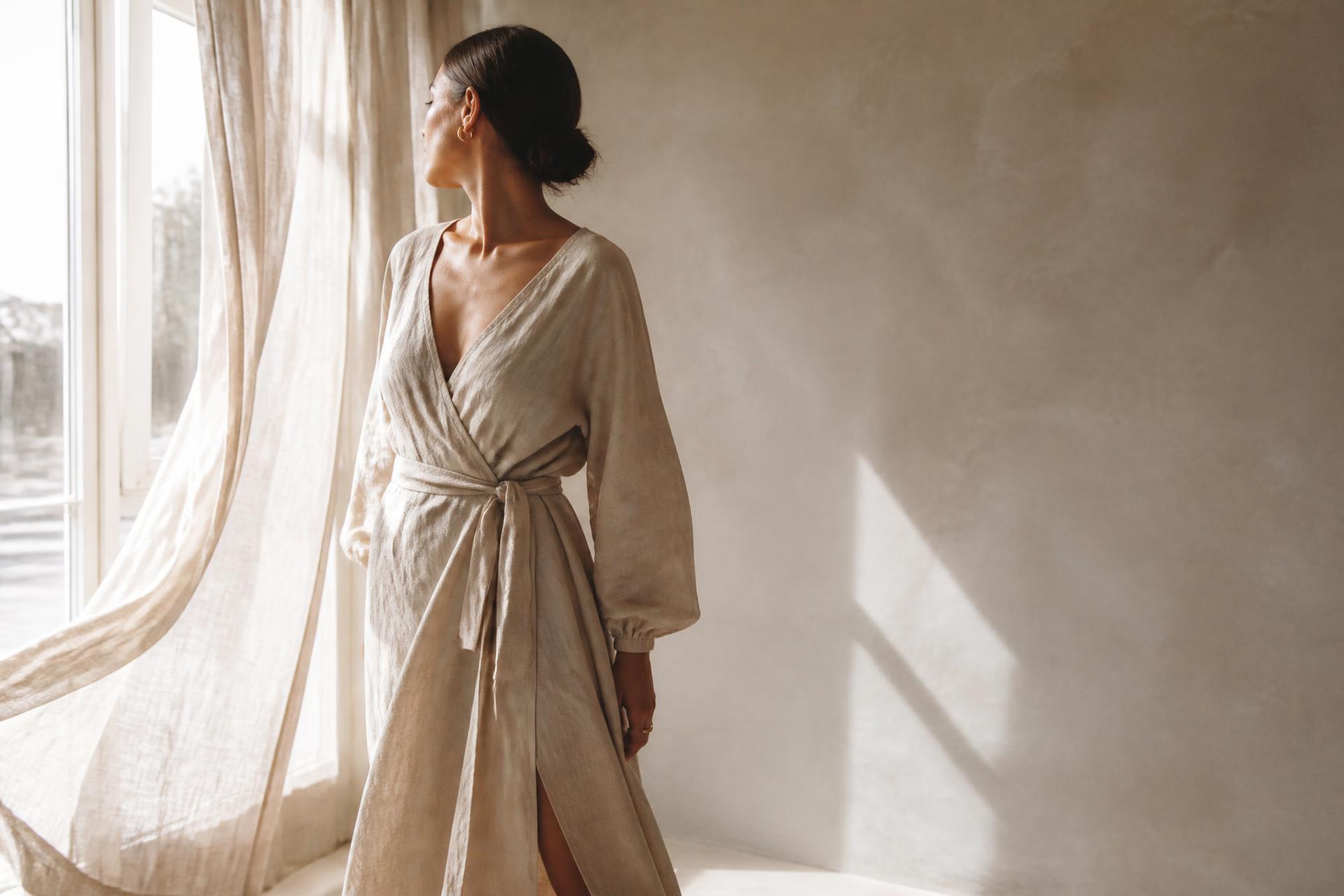 A woman in soft linen standing in warm sunlight beside a window, stepping into her next chapter